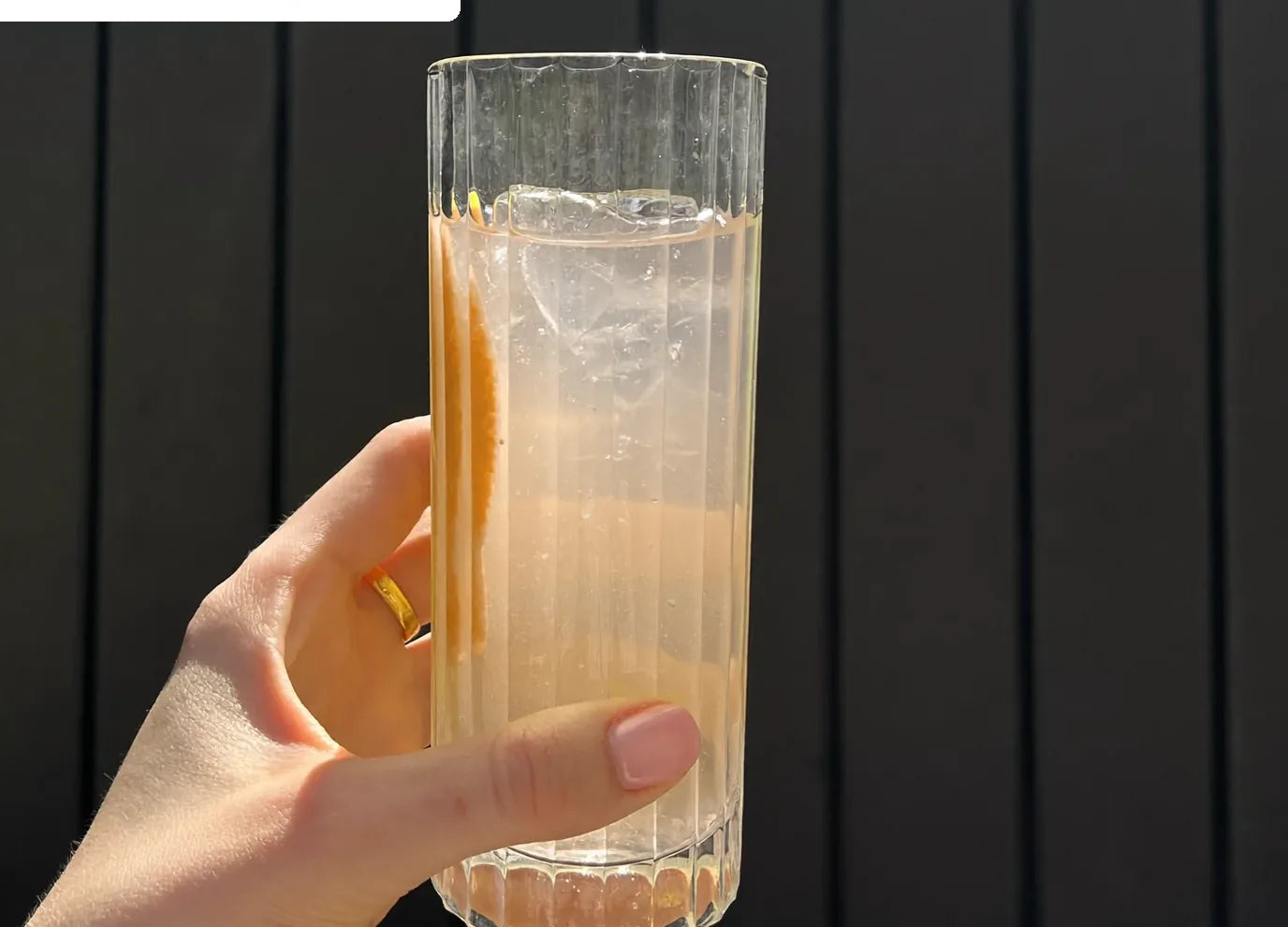 A hand holding a glass of Base 250 Tropical over ice in afternoon sun