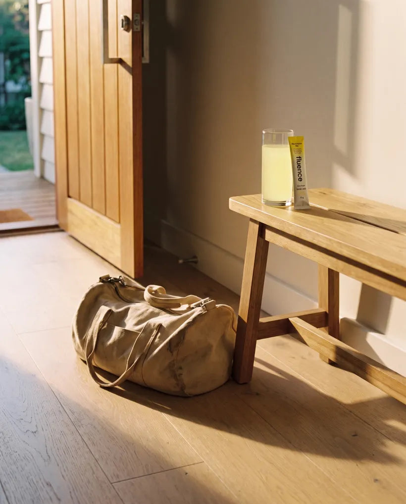 A glass of lemon Base 250 on an entryway bench beside a packed duffel bag